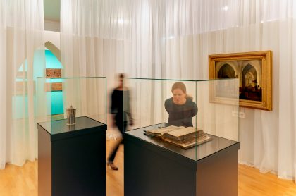 Eine Frau beugt sich in einer Ausstellung über eine Vitrine. Im Hintergrund ist unscharf eine weitere Person zu sehen.