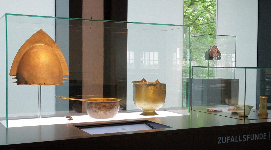 Blick in eine Vitrine. Zu sehen sind ein Helm und zwei Gefäße.