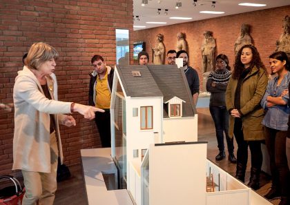 Besuchergruppe steht um das Modell eines Bremer Hauses. Eine Museumspädagogin zeigt auf das Modell.