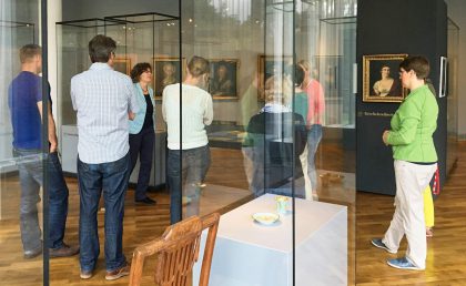 Besucherinnen und Besucher bei einer Führung. Zu sehen sind Vitrinen mit Objekten und Gemälde an den Wänden.