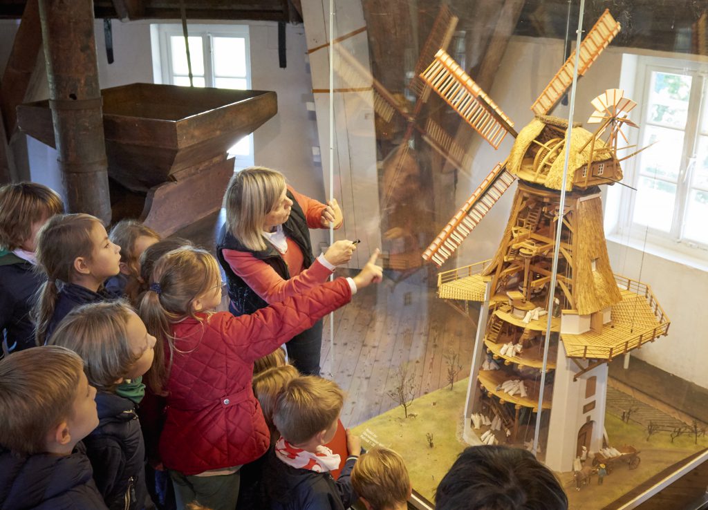 Museumspädagogin steht mit Schülerinnen und Schülern vor dem Mühlenmodell aus Holz. Sie zeigen auf etwas.