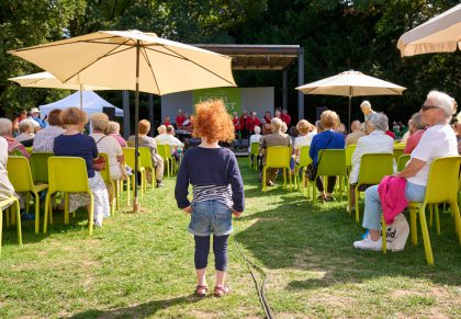 kleines Mädchen steht im Mittelgang vor der Bühne im Park, auf der gerade ein Chor singt.