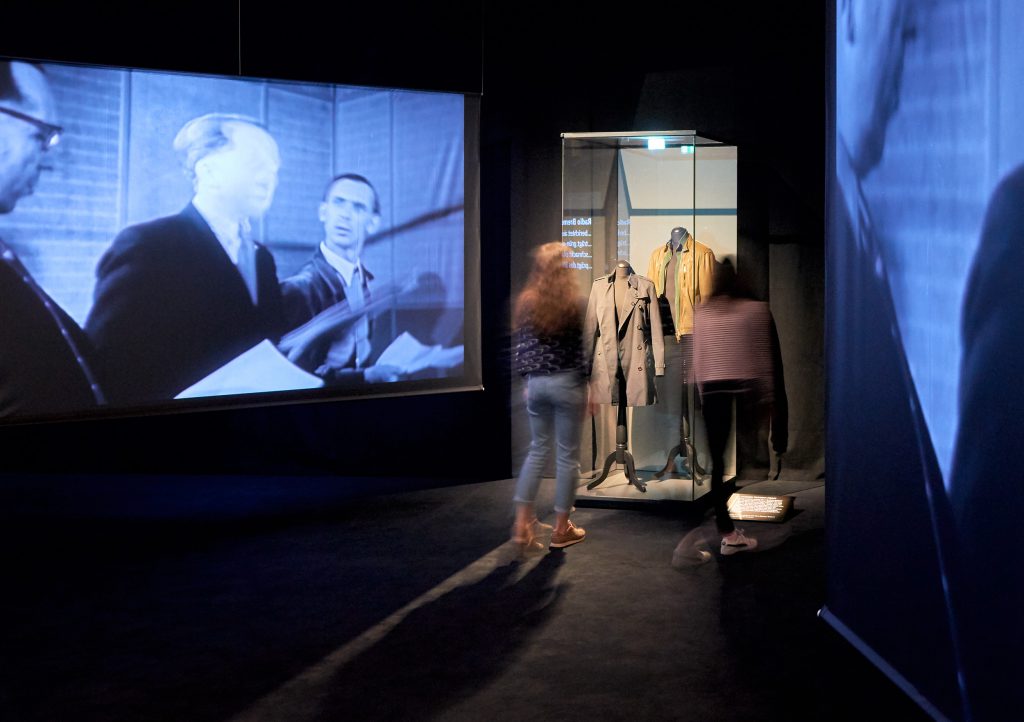 Besucher*innen schauen sich eine Vitrine in der Mediathek an. Neben ihnen wird ein Filmausschnitt auf einer großen Leinwand gezeigt.