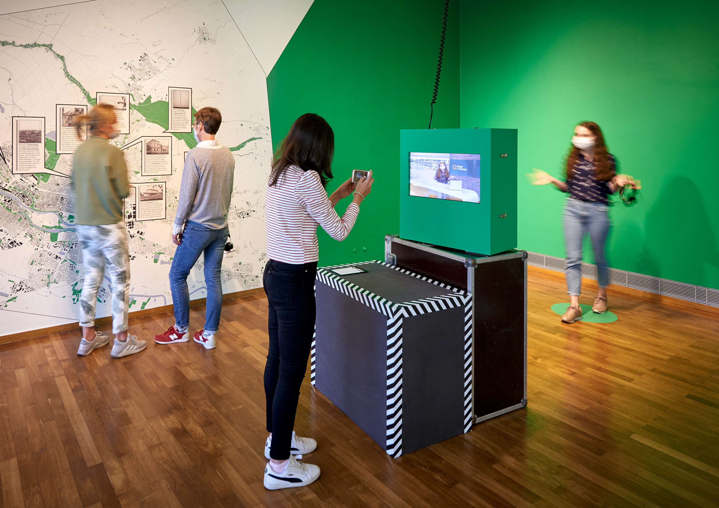 Vier Besucherinnen in der Ausstellung. Die beiden im Hintergrund sehen sich einen Stadtplan an, die beiden im Vordergrund beschäftigen sich mit dem Greenscreen vom buten un binnen-Studio. 