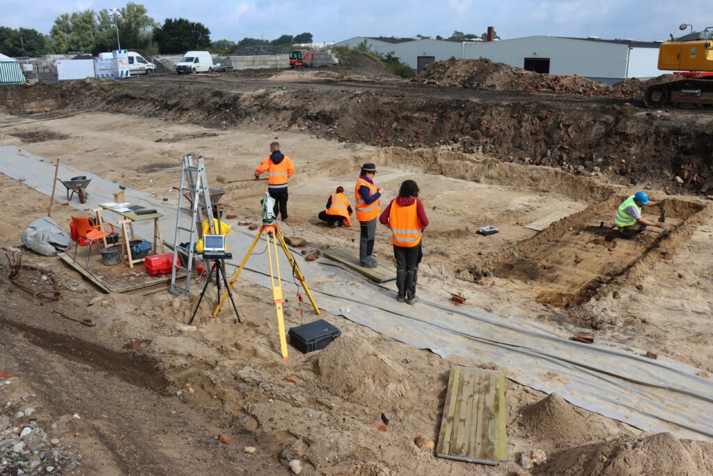 Archäologinnen und Archäologen in organgefarbenen Warnwesten auf einem Ausgrabungsfeld