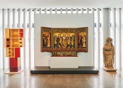 Holzschrank mit ausklappbaren Flügeln neben einem Flügelaltar.