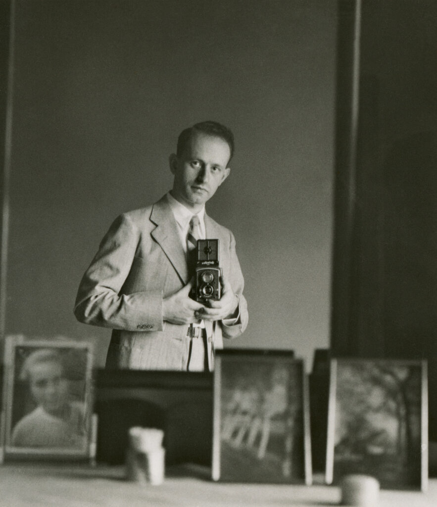 Julius Frank fotografiert sich im Spiegel. Vor ihm stehen auf einer Kommode Bilderrahmen mit Fotos von seiner Frau und Landschaftsmotiven aus seiner Heimat.