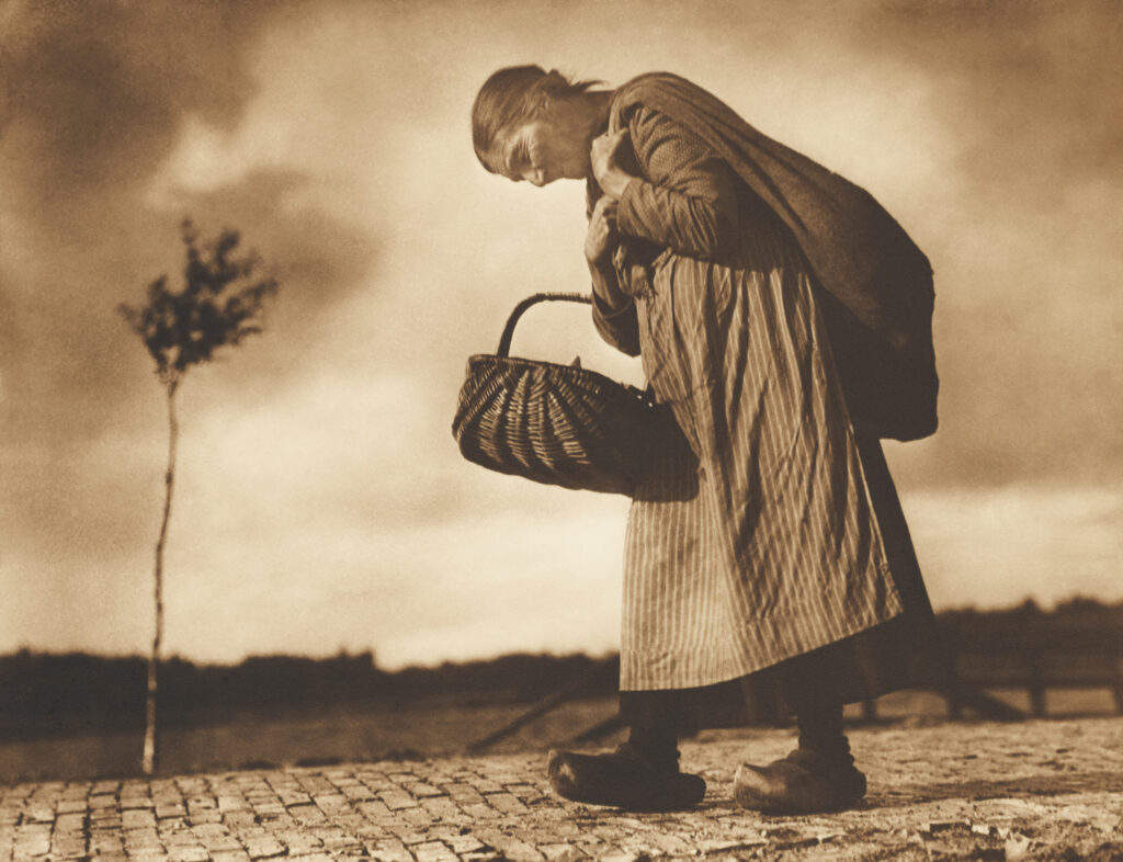 Eine alte Frau geht gebeugt einen gepflasterten Weg entlang. Sie trägt einen Korb und einen Sack über der Schulter.