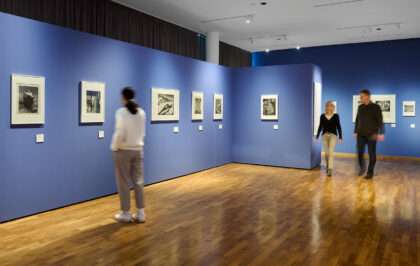 Eine Besucherin schaut sich schwarz-weiße Fotografien an einer blauen Wand an. Zwei Besucher laufen durch die Ausstellung.