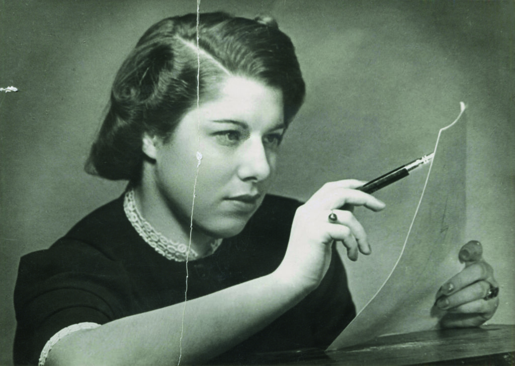 Eine junge Frau, elegant gekleidet, zeichnet mit einem Stift auf ein Blatt Papier. Es handelt sich um Franci Rabinek, die 1936 als Modedesignerin in Prag arbeitete. 