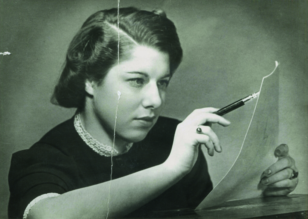 Eine junge Frau, elegant gekleidet, zeichnet mit einem Stift auf ein Blatt Papier. Es handelt sich um Franci Rabinek, die 1936 als Modedesignerin in Prag arbeitete.