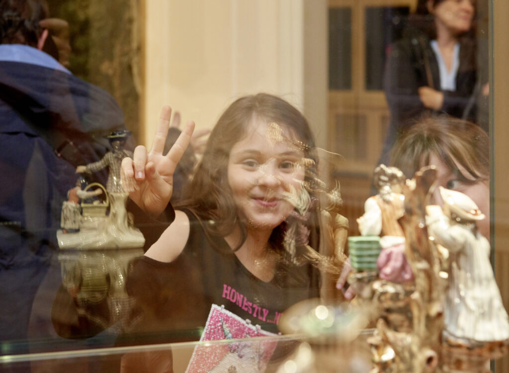 Mädchen schaut durch eine Vitrine mit Porzellanfiguren und macht mit den Fingern ein Victory-Zeichen. 