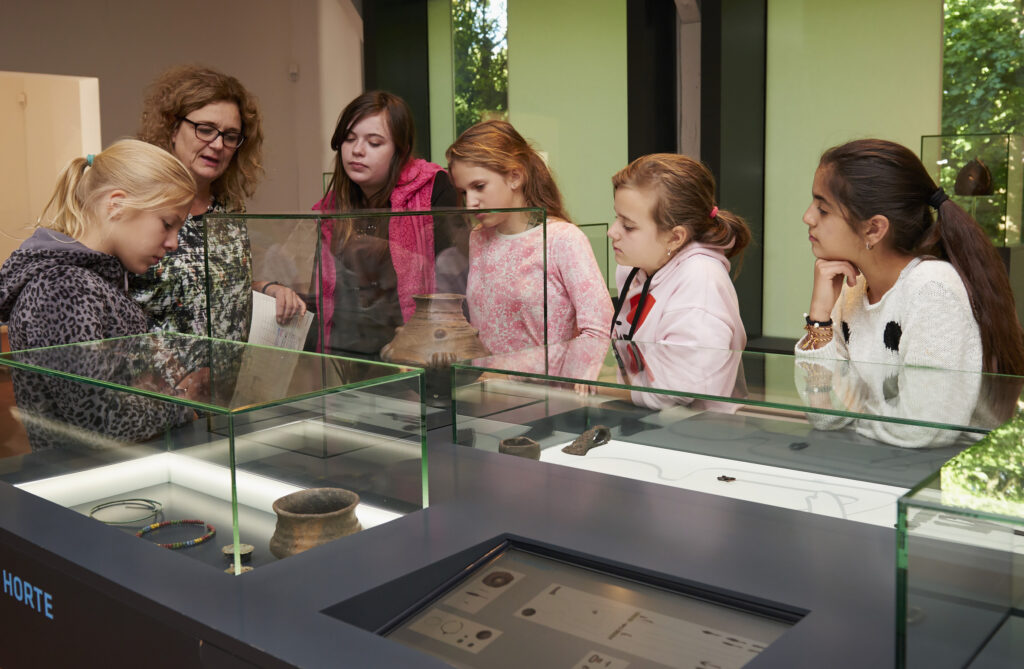 Kinder und eine Museumspädagogin schauen sich archäologische Objekte in einer Vitrine an.