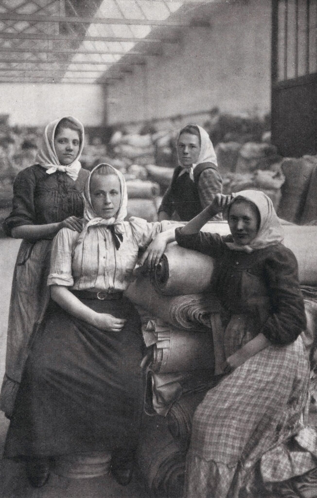 Vier Frauen mit Röcken, Blusen und Kopftüchern sitzen in einer Lagerhalle auf Stoffrollen.