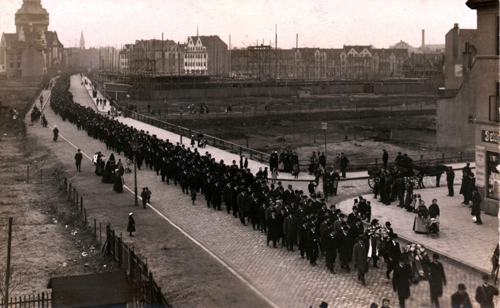 schwarz-weiß Foto eines langen Zuges von Menschen in schwarzer Kleidung durch einen Straßenzug. Die ersten Menschen tragen zwei große Blumenkränze. Einige Passanten schauen am Straßenrand zu.
