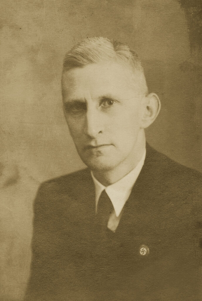 schwarz-weiß-Foto eines mittelalten Mannes. Er trägt die Haare akkurat geschnitten und einen Anzug. Auf der linken Brust ist ein Parteiabzeichen der NSDAP zu sehen.