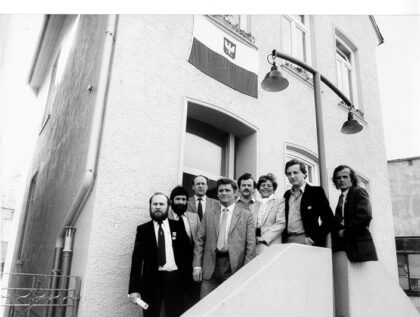 Sieben Männer und eine Frau in Anzügen stehen auf einer Treppe vor einem Haus. Oben am Haus hängt die polnische Flagge.