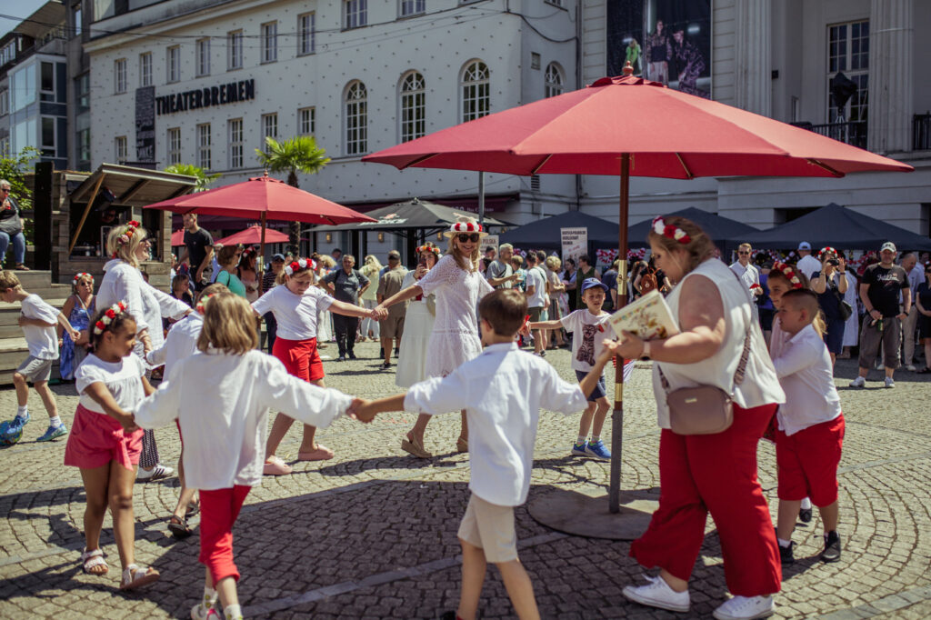 Frauen und Kinder tanzen in einem Reigen vor dem Bremer Theater. Sie sind rot und weiß gekleidet.