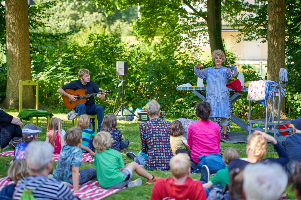 Aufführung von "Mensch Puppe" im Park. Zu sehen sind eine Schauspielerin und ein Gitarrenspieler. Im Vordergrund sitzen Kinder auf dem Rasen.